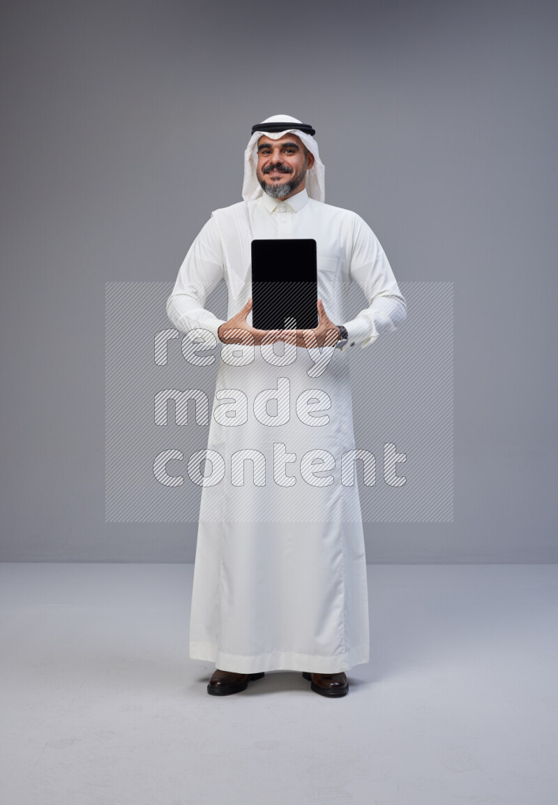 Saudi man Wearing Thob and white Shomag standing showing tablet to camera on Gray background