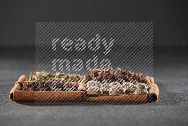 4 مربعات من عيدان القرفة ممتلئة بحب الهيل واليانسون النجمي وجوزة الطيب والقرنفل على أرضية سوداء