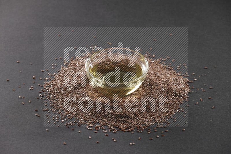 A glass bowl full of flaxseeds oil surrounded by the seeds on a black flooring