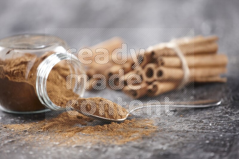 Herbal glass jar full cinnamon powder flipped and a metal spoon full of powder, cinnamon sticks stacked and bounded in the back on textured black background in different angles