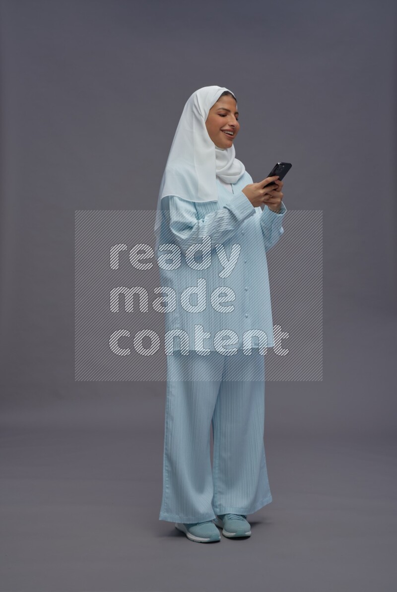 Saudi woman wearing hijab clothes standing texting on phone on gray background