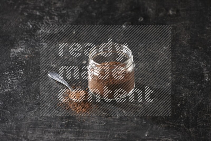 A glass jar full of cloves powder with a metal spoon on a textured black flooring
