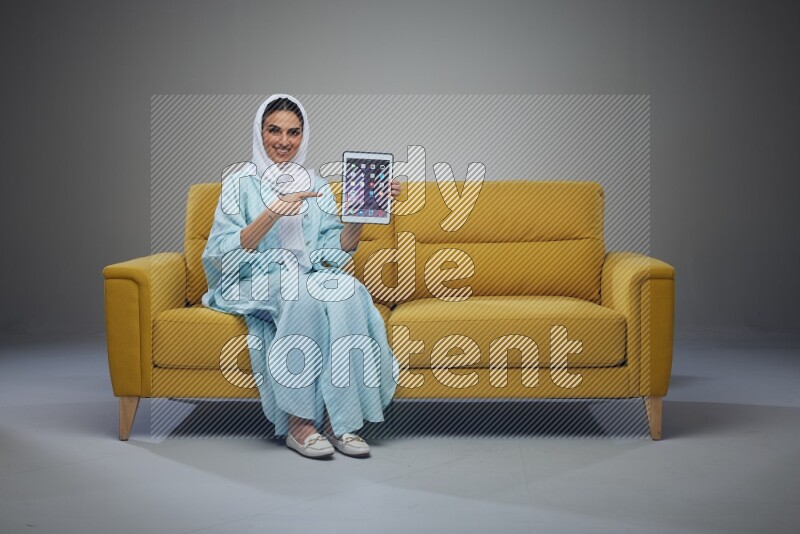A Saudi woman wearing a light blue Abaya and white head scarf sitting on a yellow sofa and showing her tablet's screen while pointing to it eye level on a grey background