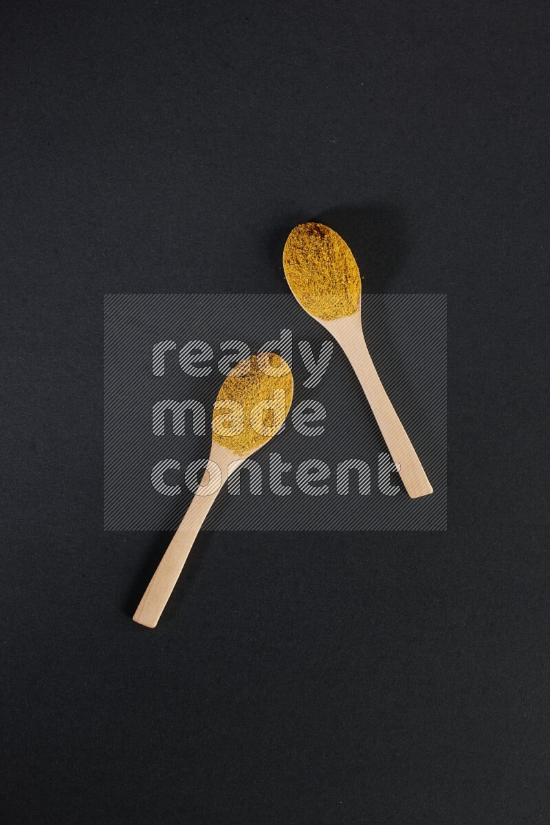 2 wooden spoons full of turmeric powder on black flooring
