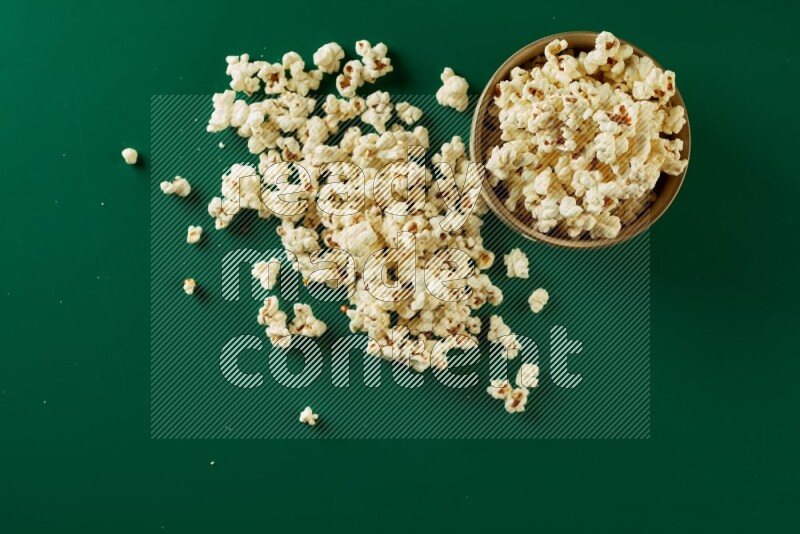 An off white ceramic bowl full of popcorn with popcorn beside it on a green background in a top view shot