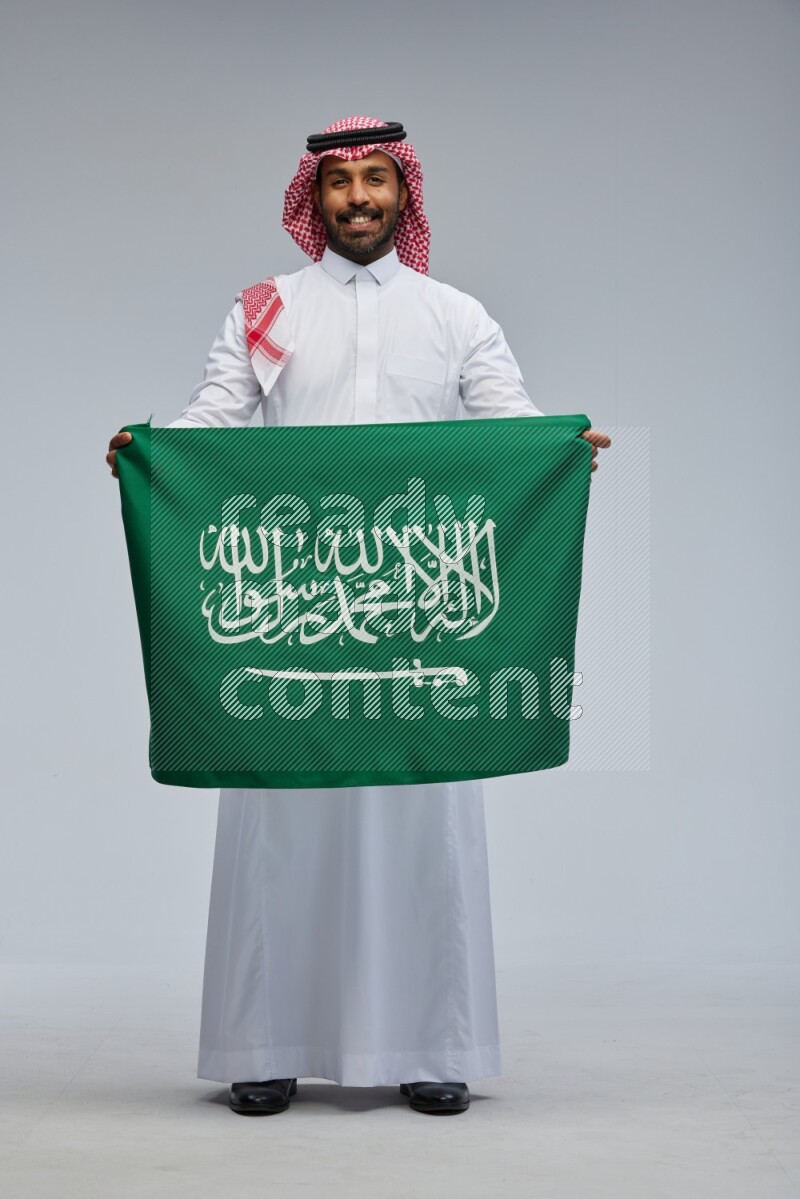 Saudi man Wearing Thob and shomag standing holding Saudi flag on Gray background