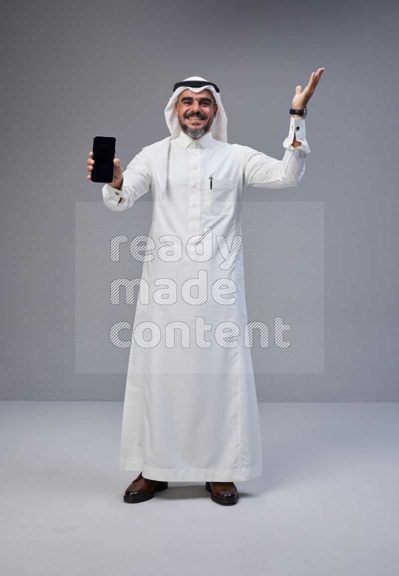 Saudi man Wearing Thob and white Shomag standing showing phone to camera on Gray background