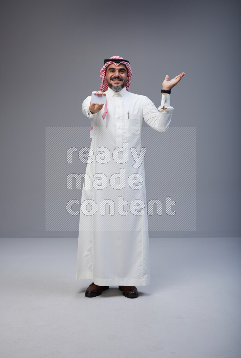 Saudi man Wearing Thob and red Shomag standing holding ATM card on Gray background