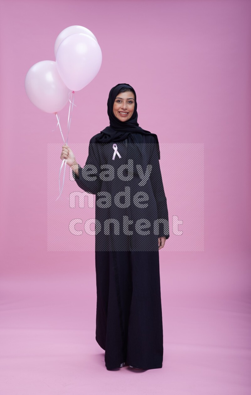 Saudi woman wearing pink ribbon on Abaya standing holding balloons on plain pink background