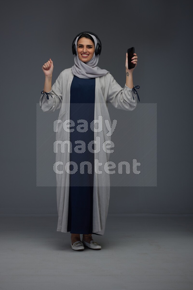 A Saudi woman wearing a light gray Abaya and head scarf standing and listening to music on a grey background
