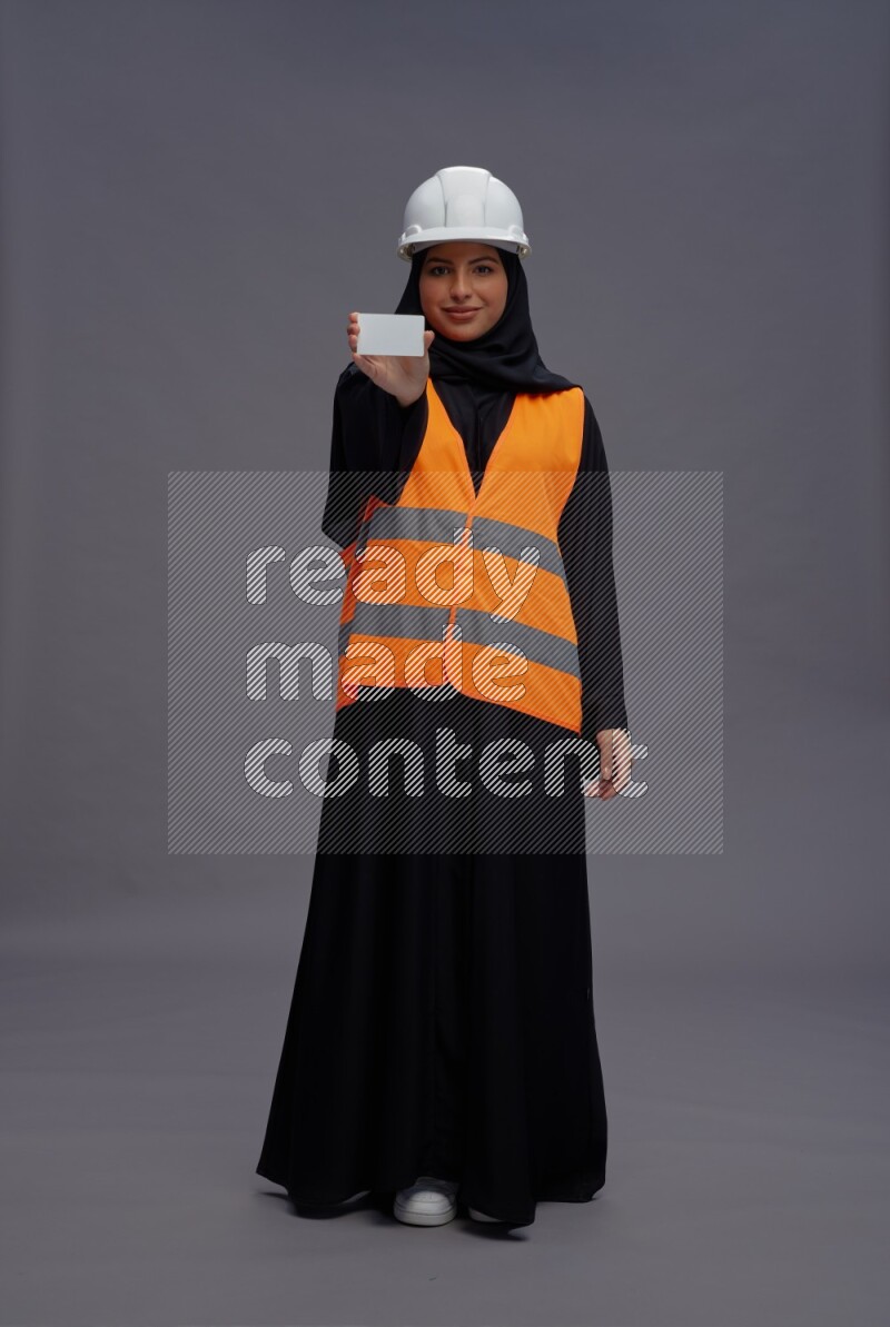 Saudi woman wearing Abaya with engineer vest standing holding ATM card on gray background