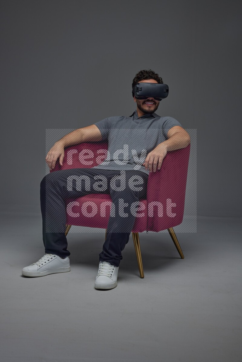 A man wearing casual and wearing VR while sitting on a burgundy chair eye level on a gray background
