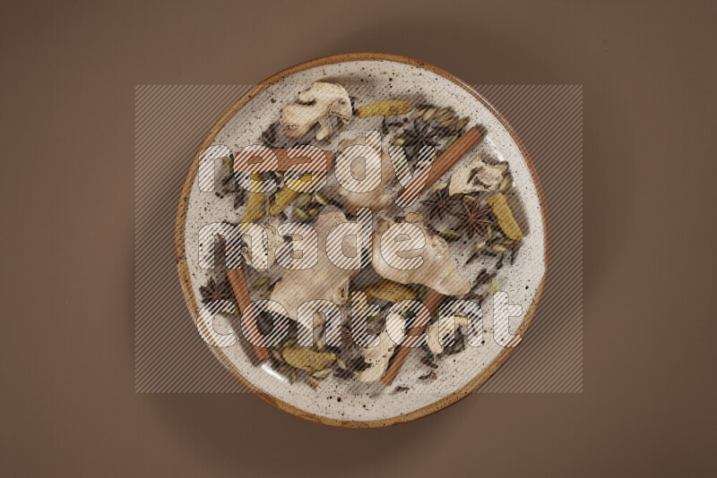 An assorted whole spices on a flat beige plate including fresh and dried ginger, cinnamon sticks, cardamom, turmeric fingers, cloves, star anise on a beige background