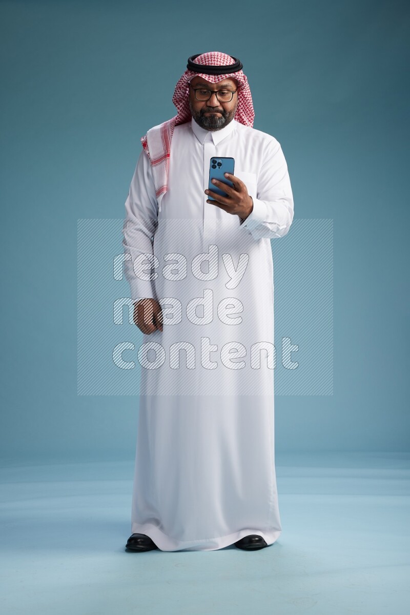 Saudi Man with shimag Standing texting on phone on blue background