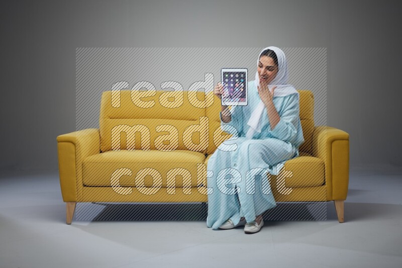 A Saudi woman wearing a light blue Abaya and white head scarf sitting on a yellow sofa and showing her tablet's screen while pointing to it eye level on a grey background