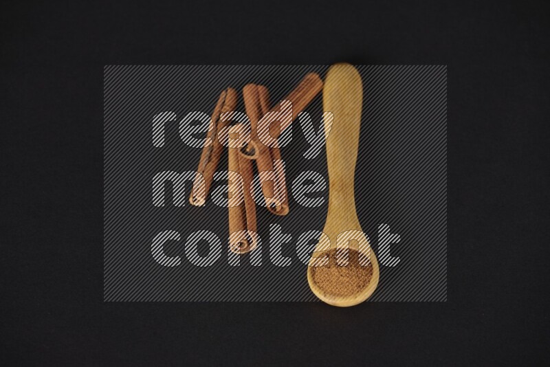 Cinnamon powder in a wooden spoon with cinnamon sticks beside it on black background