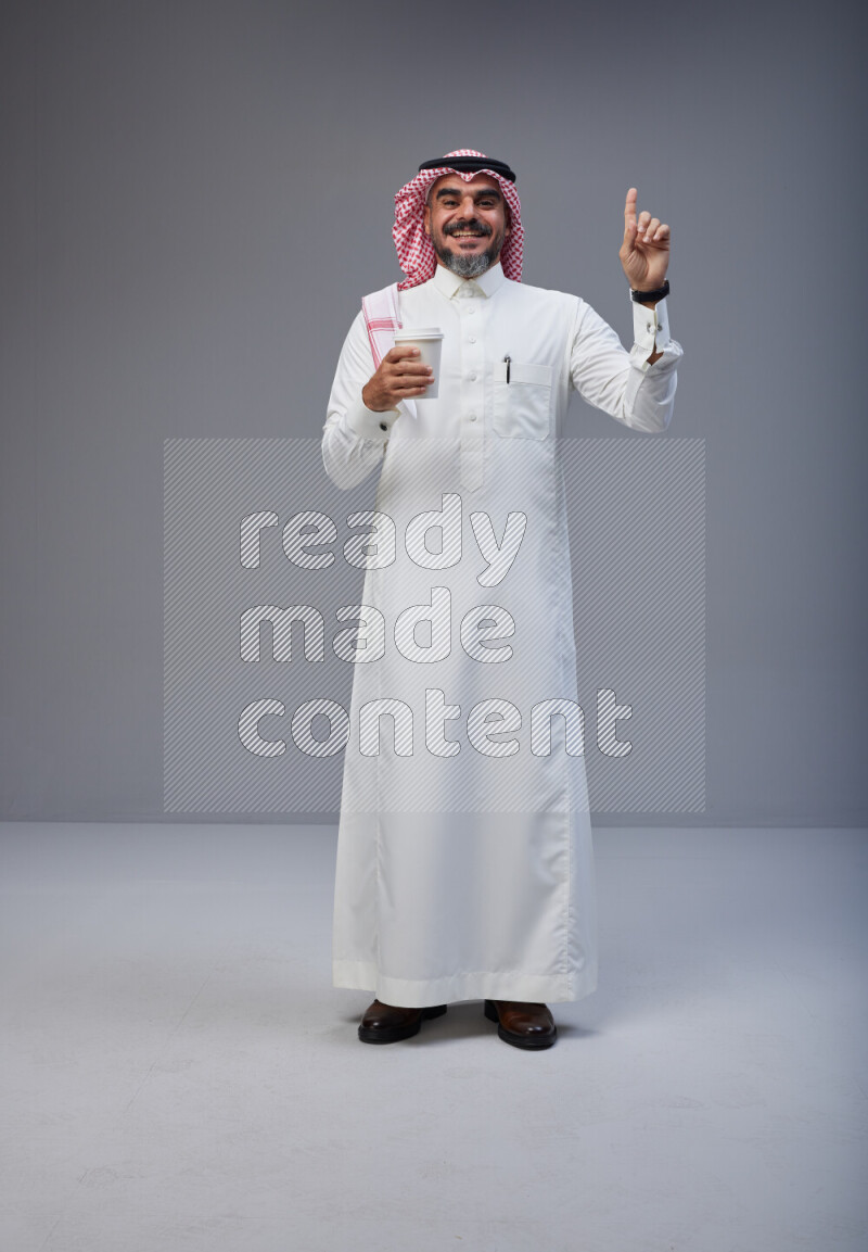Saudi man Wearing Thob and red Shomag standing holding paper cup on Gray background