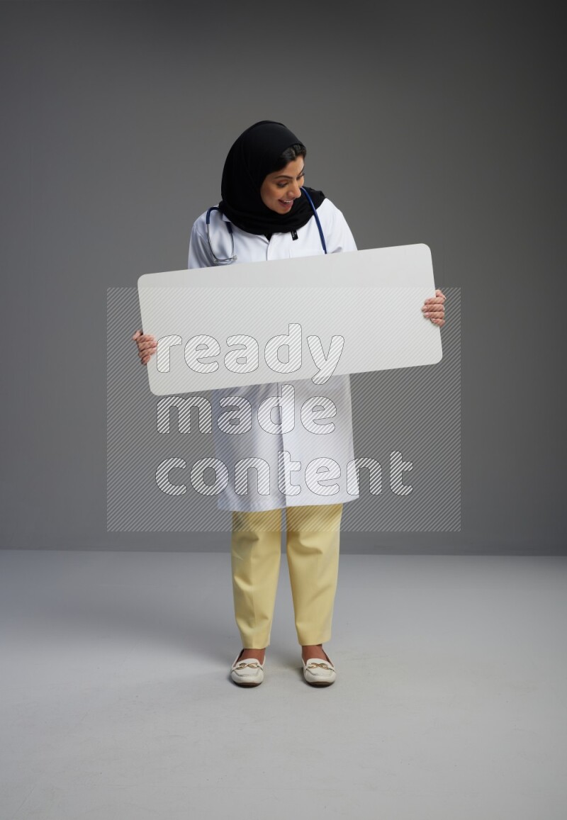 Saudi woman wearing lab coat with stethoscope standing holding board on Gray background