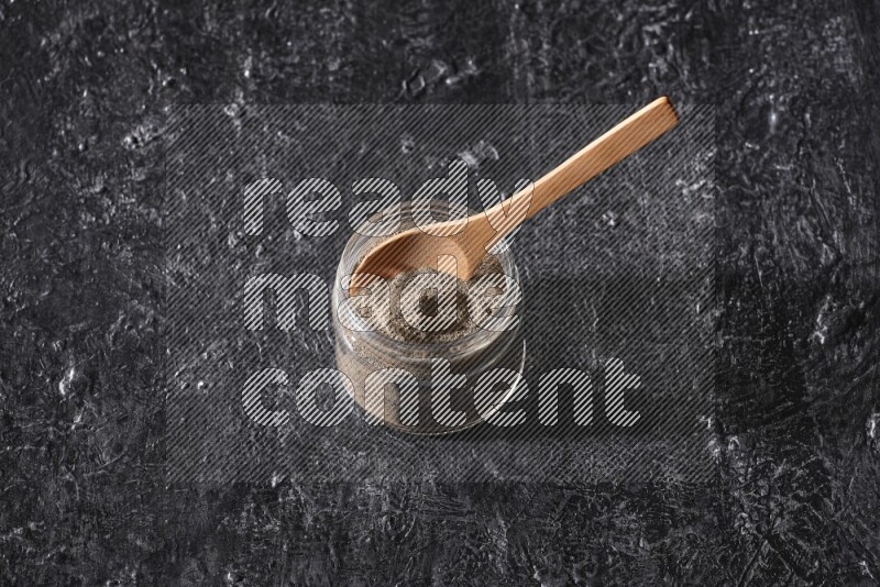 A glass jar full of black pepper powder and a wooden spoon on a textured black flooring