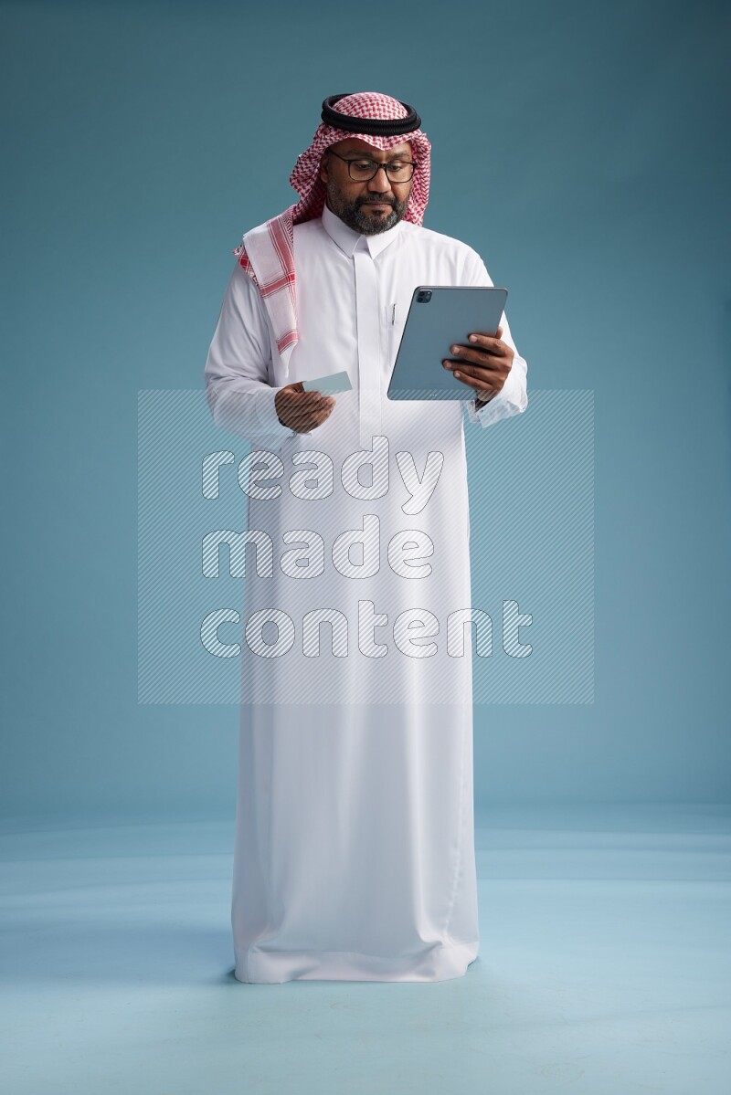 Saudi Man with shimag Standing holding ATM card while working on tablet on blue background
