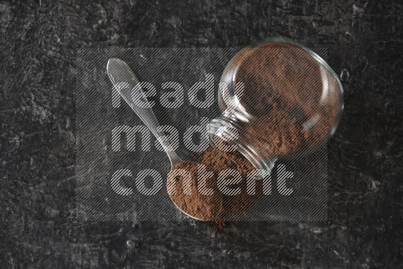 A flipped glass spice jar and a metal spoon full of cloves powder on textured black flooring