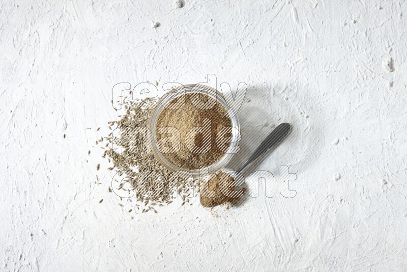 A glass bowl and metal spoon full of cumin powder and cumin seeds underneath it on textured white flooring