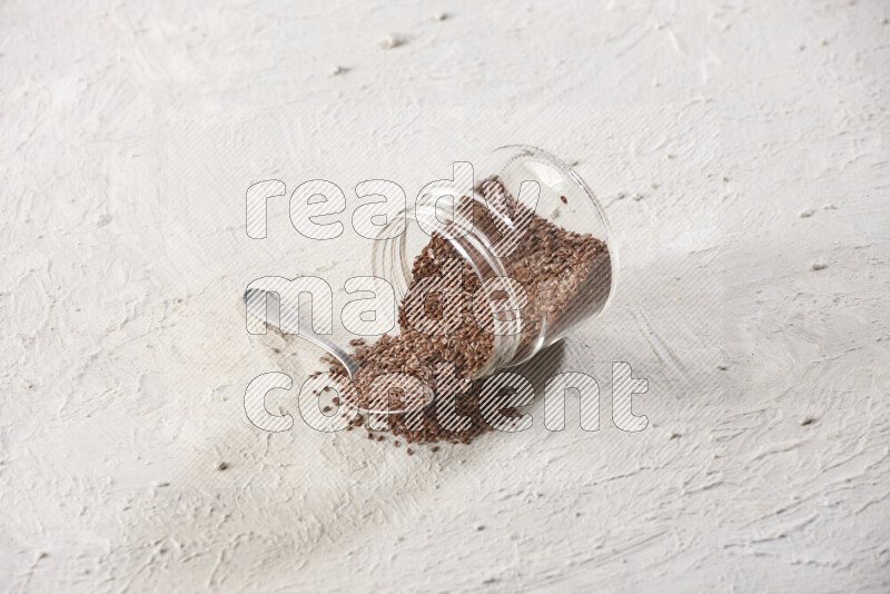 A glass spice jar full of flax seeds flipped with a metal spoon full of the seeds on a textured white flooring