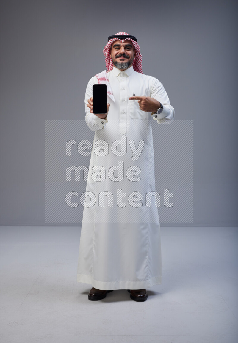 Saudi man Wearing Thob and red Shomag standing showing phone to camera on Gray background