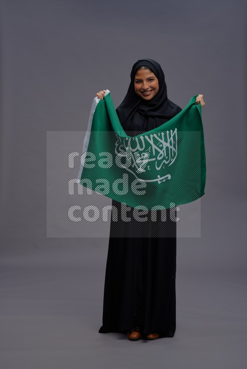 Saudi woman wearing Abaya standing holding Saudi flag on gray background