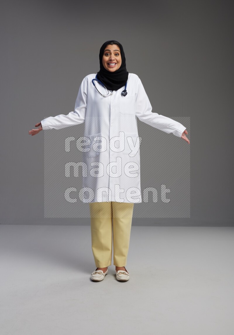 Saudi woman wearing lab coat with stethoscope standing interacting with the camera on Gray background