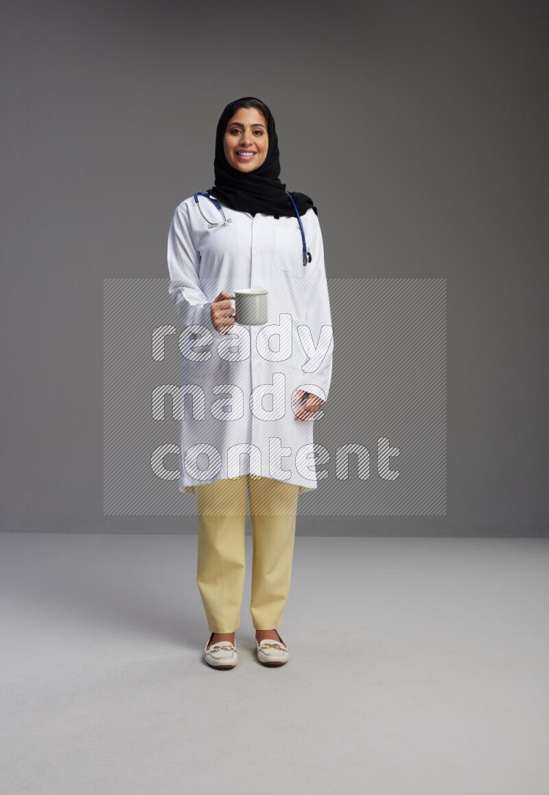 Saudi woman wearing lab coat with stethoscope standing holding mug on Gray background