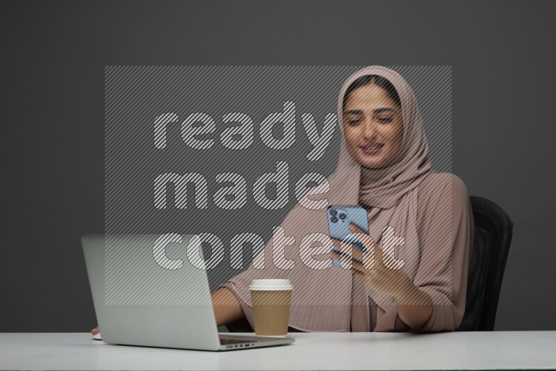 A Saudi woman Sitting on her desk Texting on a Gray Background wearing Brown Abaya with Hijab