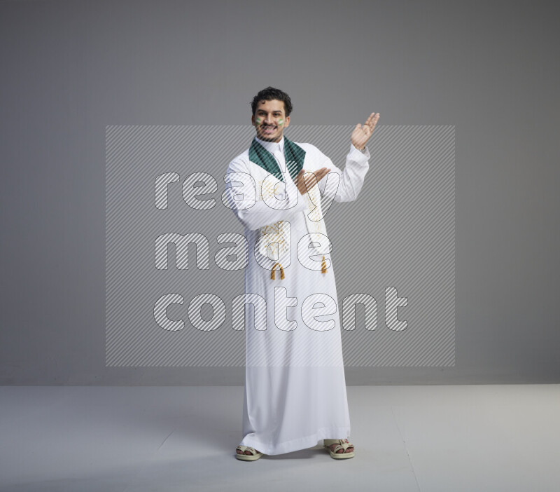 A Saudi man standing wearing thob and Saudi flag scarf with face painting on gray background