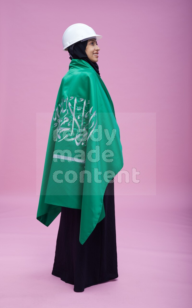 Saudi woman wearing Abaya with engineer vest and helmet standing holding Saudi flag on pink background