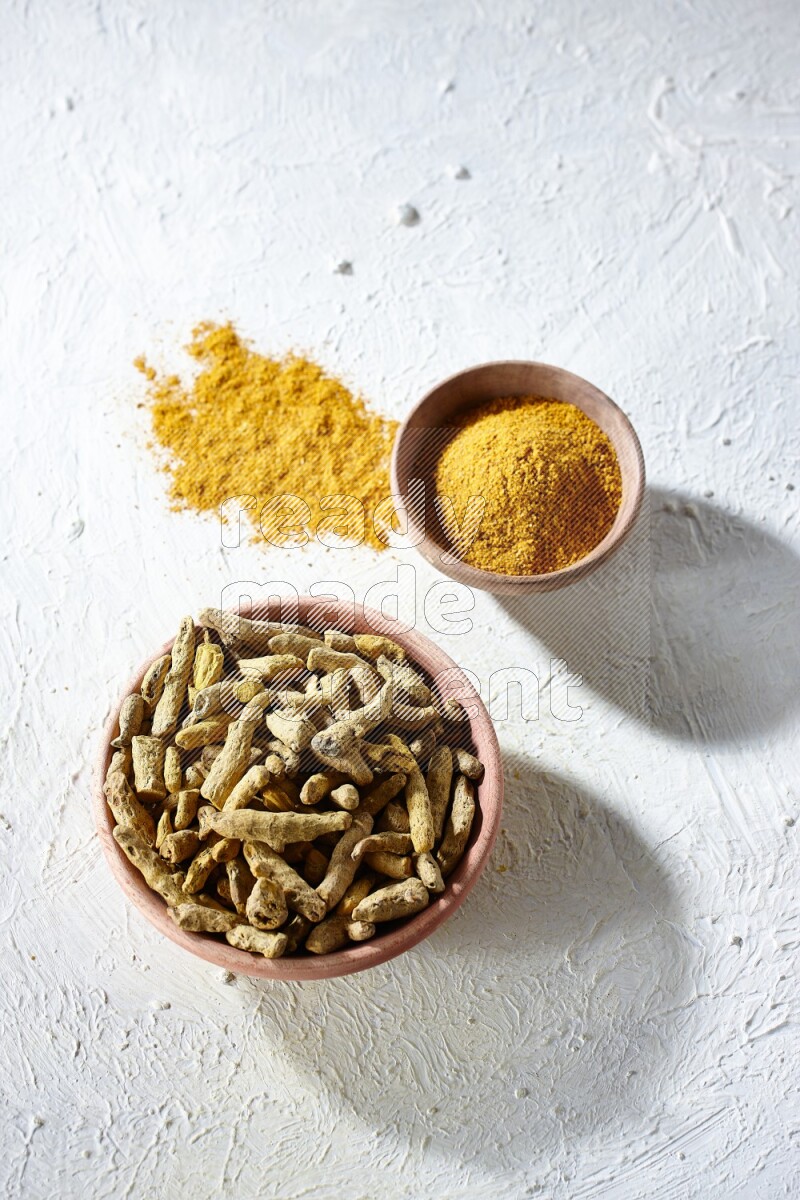 2 wooden bowls, one full of turmeric powder and the other full of dried turmeric whole fingers on a textured white flooring