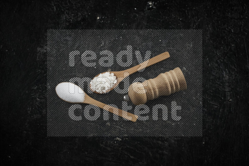2 wooden spoons filled with fine and coarse salt with wooden grinder beside them on black background