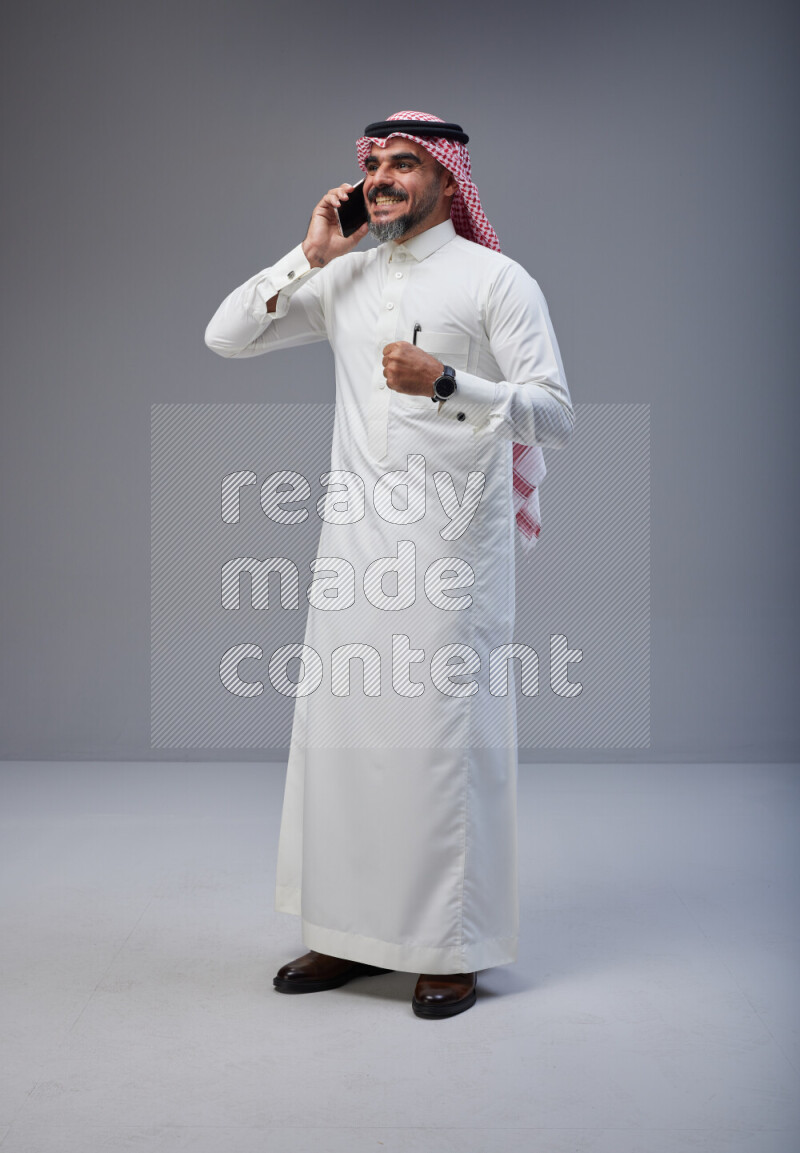 Saudi man Wearing Thob and red Shomag standing talking on phone on Gray background