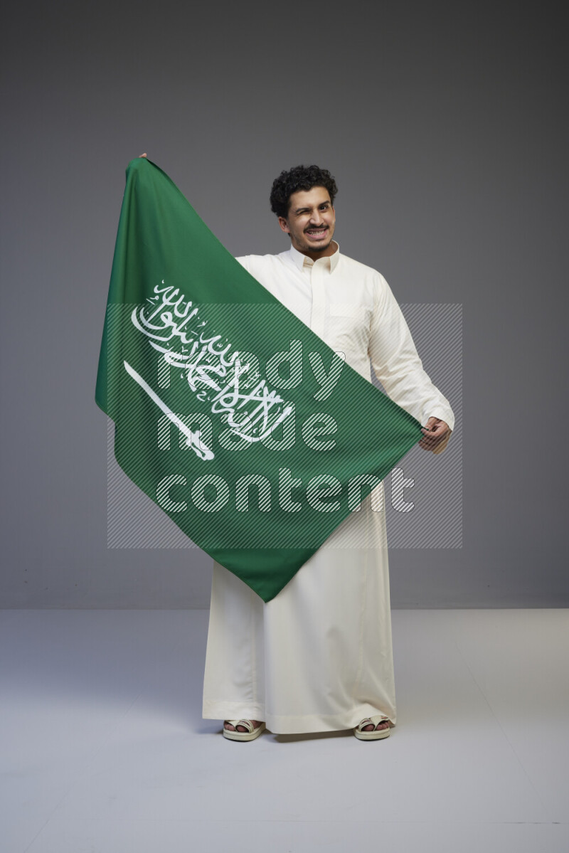 A Saudi man standing wearing thob holding big Saudi flag on gray background