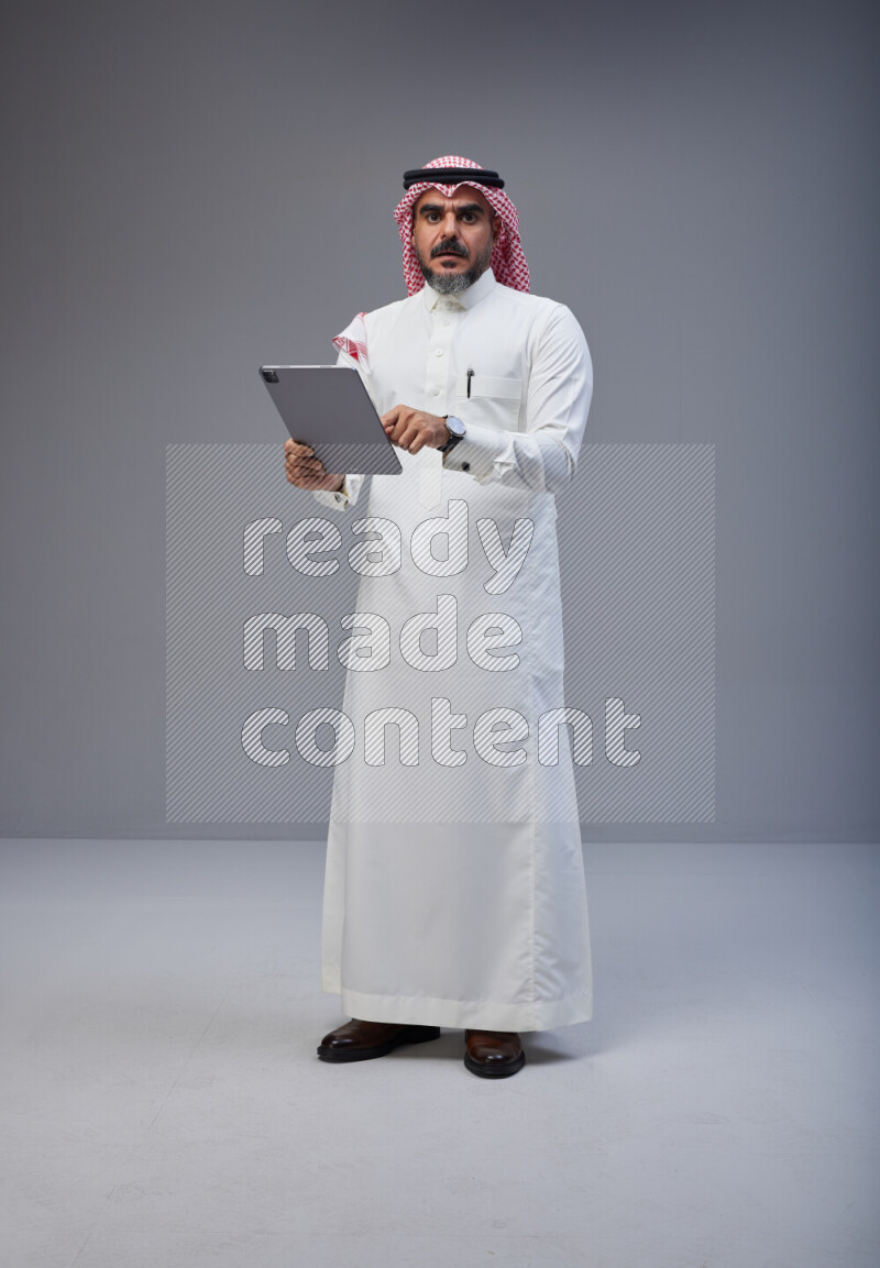 Saudi man Wearing Thob and red Shomag standing working on tablet on Gray background