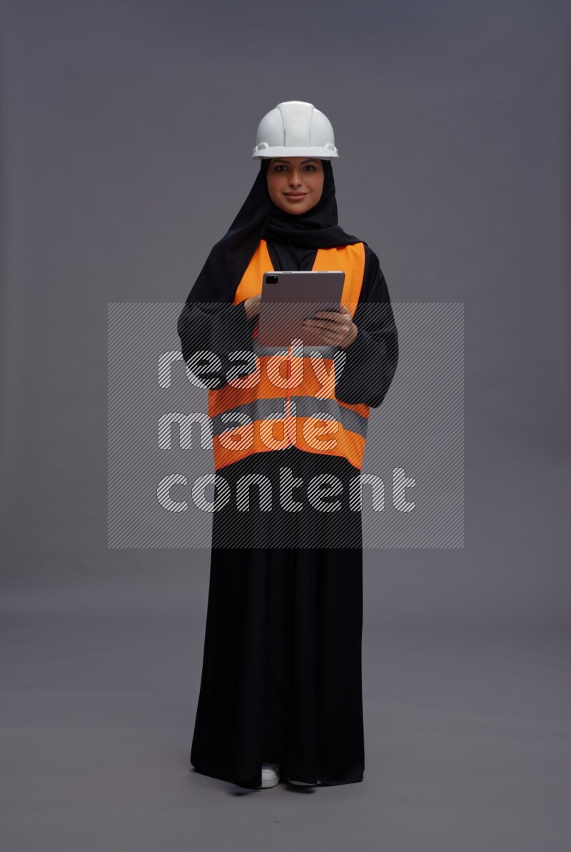 Saudi woman wearing Abaya with engineer vest standing working on tablet on gray background