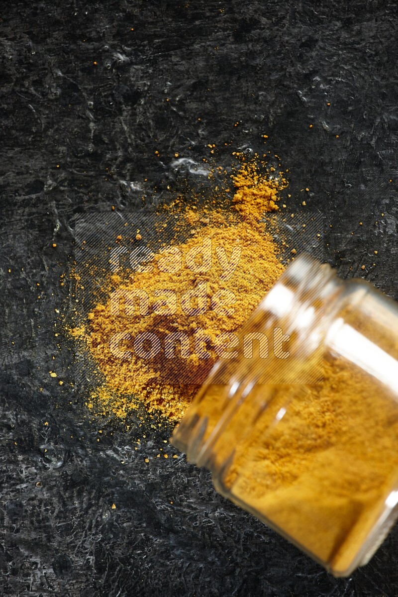 A flipped glass jar full of turmeric powder and powder spilled out of it on a textured black flooring