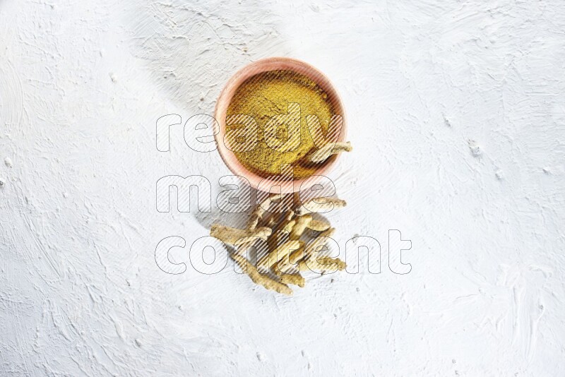 A wooden bowl full of turmeric powder and dried turmeric whole fingers beside it on textured white flooring