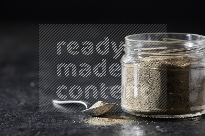 A glass jar and a metal spoon full of white pepper powder on textured black flooring