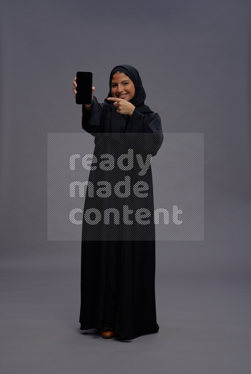 Saudi woman wearing Abaya standing showing phone to camera on gray background