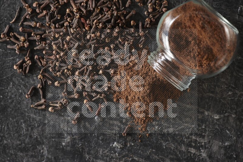 A flipped glass spice jar full of cloves powder and powder came out of it with cloves spread on textured black flooring
