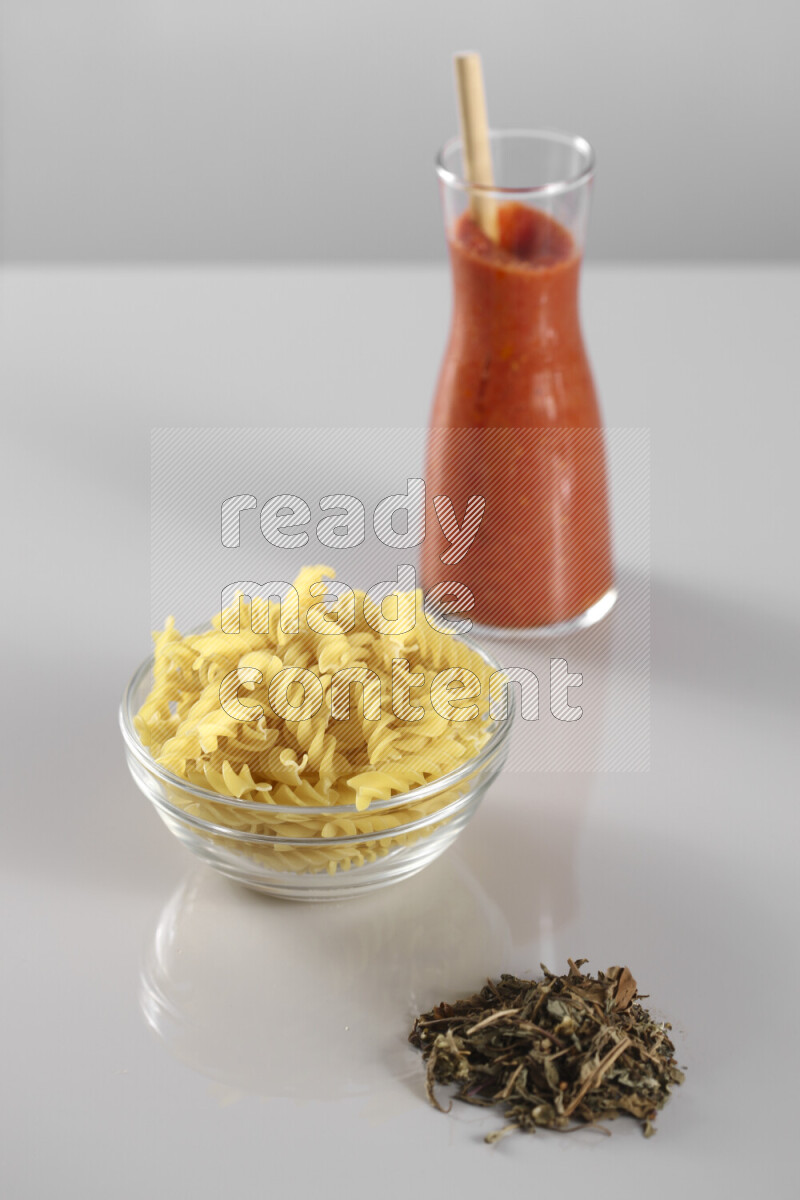 Raw pasta with tomatoe pasta with different ingredients such as cherry tomatoes, basil, garlic, bay laurel, cardamom, white pepper, black pepper, red chilis and wheat stalks on light grey background