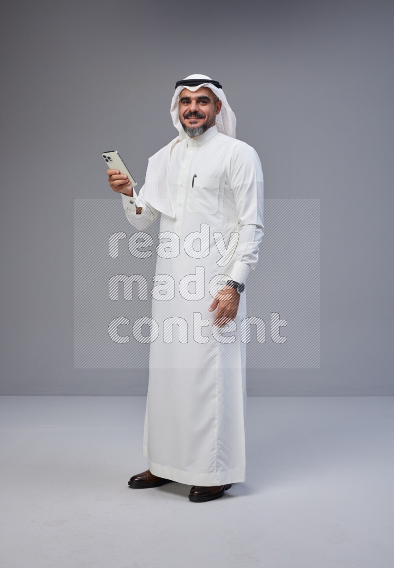 Saudi man Wearing Thob and white Shomag standing texting on phone on Gray background