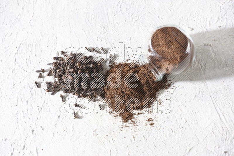 A glass spice jar full of cloves powder flipped and powder came out of it with cloves spread on textured white flooring