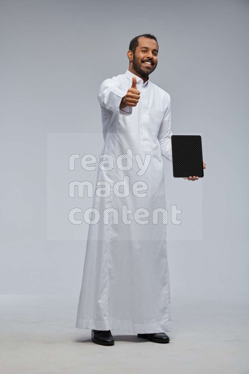 Saudi man wearing Thob standing showing tablet to camera on Gray background