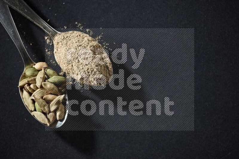 2 Metal spoons full of cardamom powder and cardamom seeds on black flooring